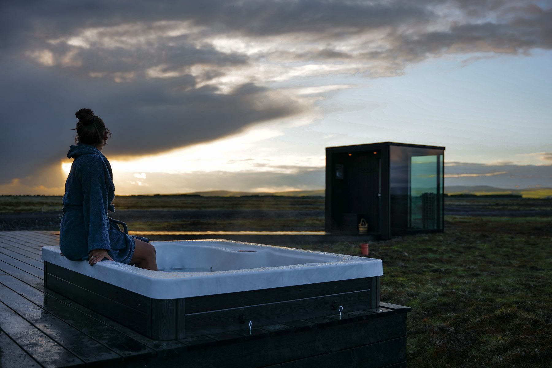 Guest sitting on the edge of the private hot tub at sunset, glass lodge behind, Harmony Seljalandsfoss Iceland